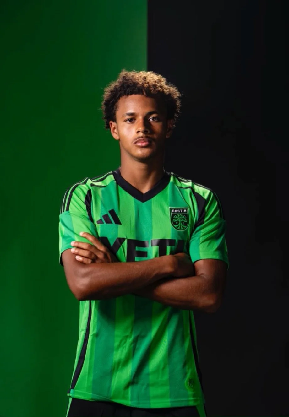 A young male soccer player with curly hair, wearing a green and black Austin FC jersey, standing against a split green and black background, with arms crossed and a serious expression.