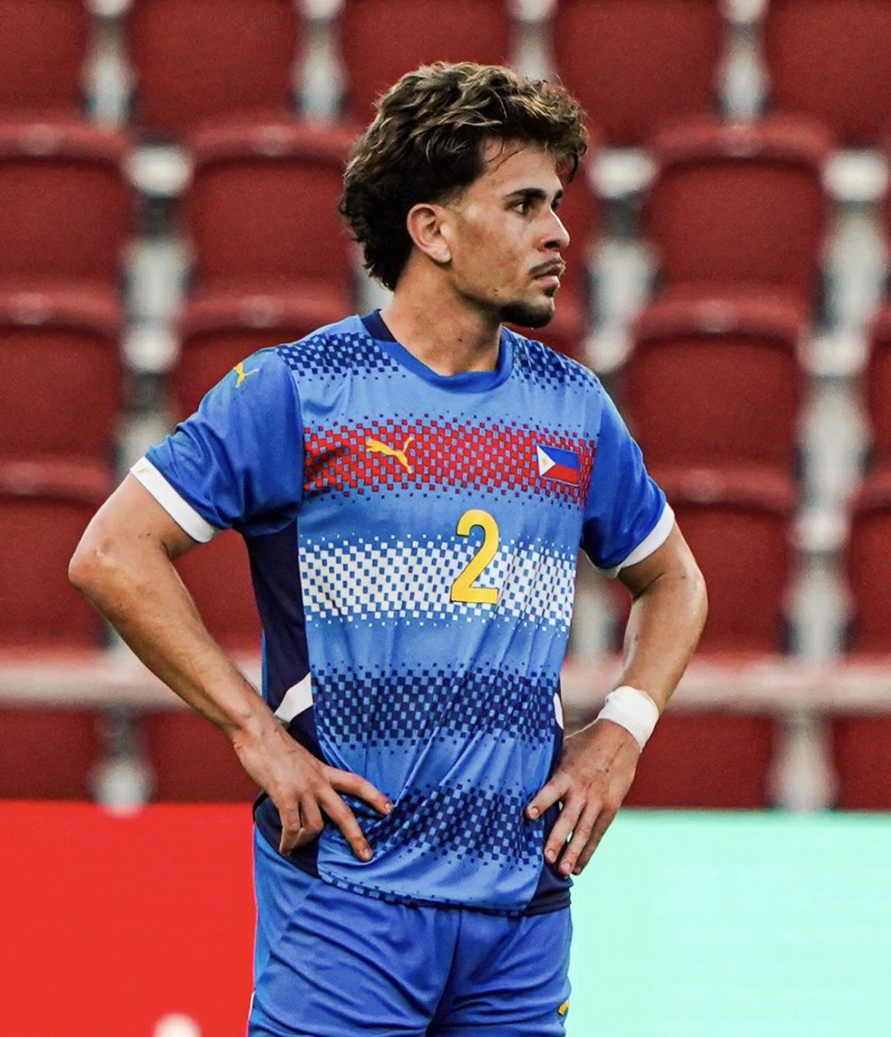 A soccer player in a blue jersey with the number 2, standing on the field with hands on hips, looking to the side, with red stadium seats in the background.