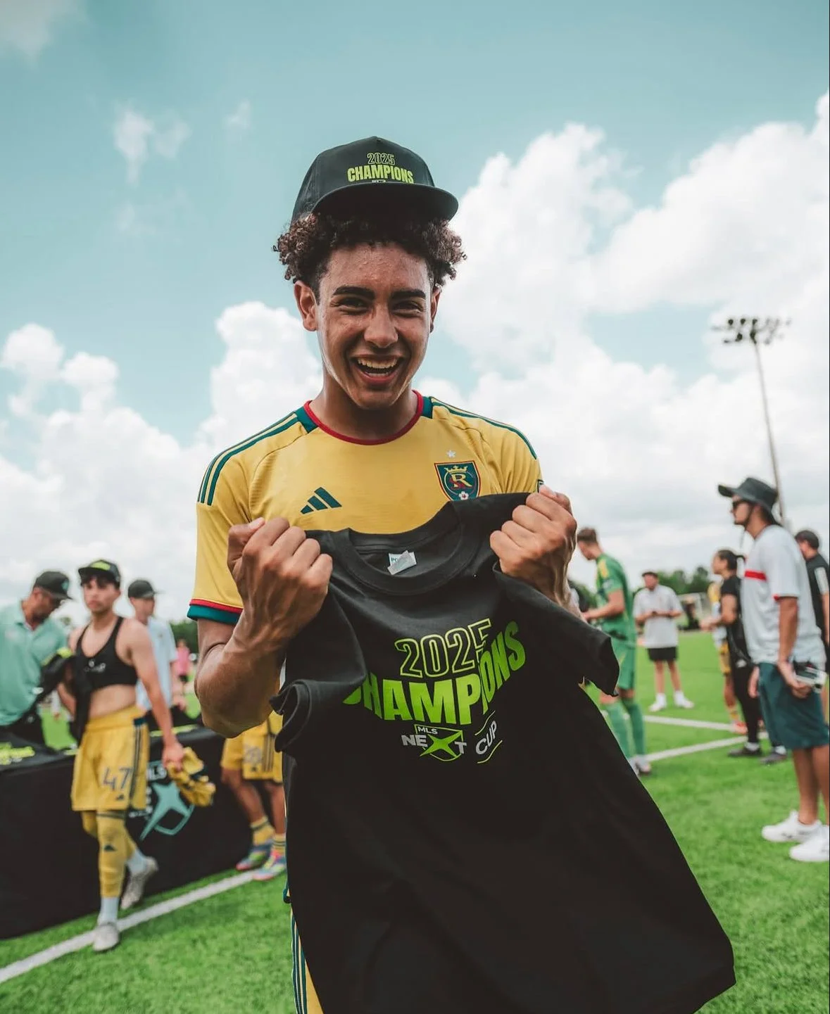Young man in yellow soccer jersey celebrating with a black championship shirt in a sports field, with other people in the background.
