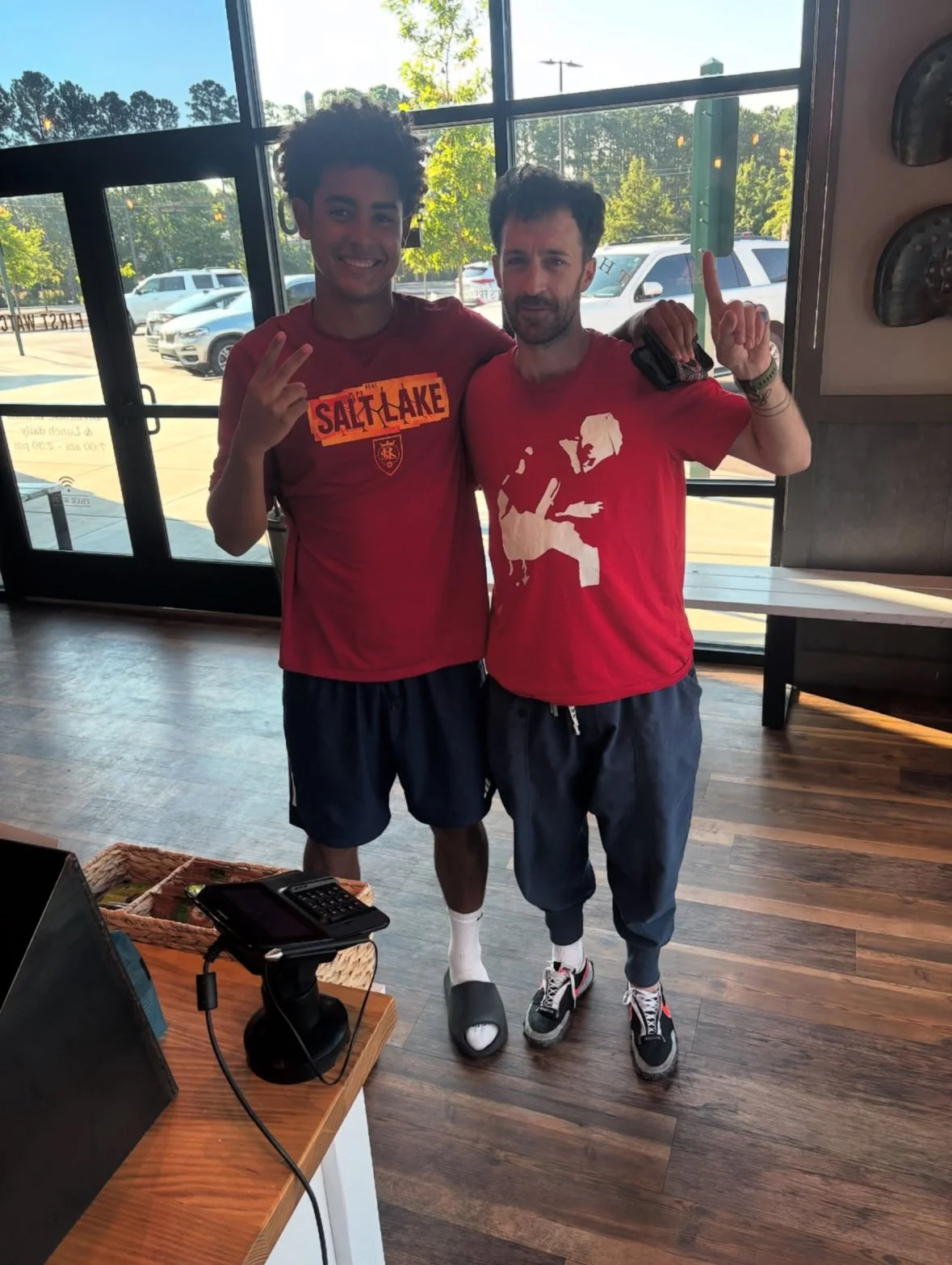 Two young men standing inside a restaurant near large windows, smiling and posing for a photo. One is wearing a red shirt with a Salt Lake City logo and the other is wearing a red graphic t-shirt. They are making peace signs and pointing up. There are cars visible outside through the windows.
