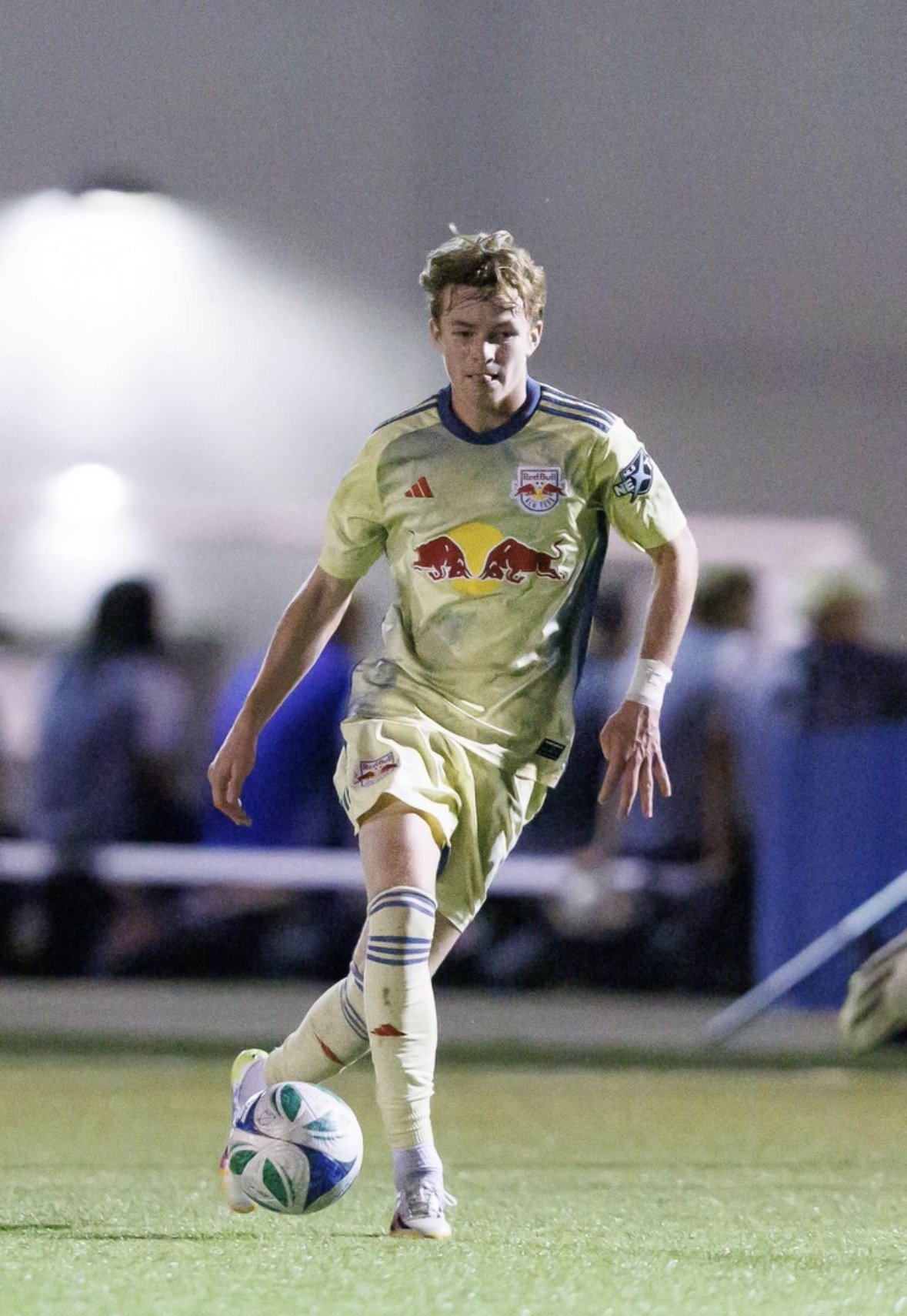 A young male soccer player in a yellow uniform is running on the field, kicking a soccer ball during a game at night.