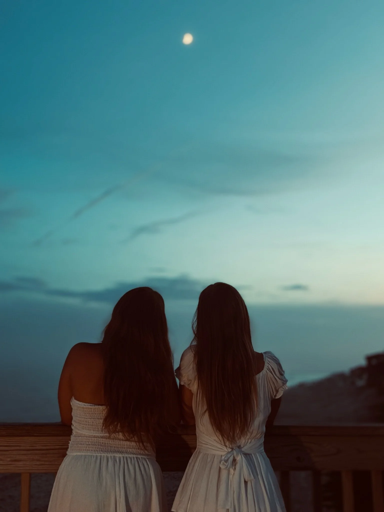 Two women in white dresses sitting on a wooden railing, watching the sky with the moon visible, during dusk or dawn.