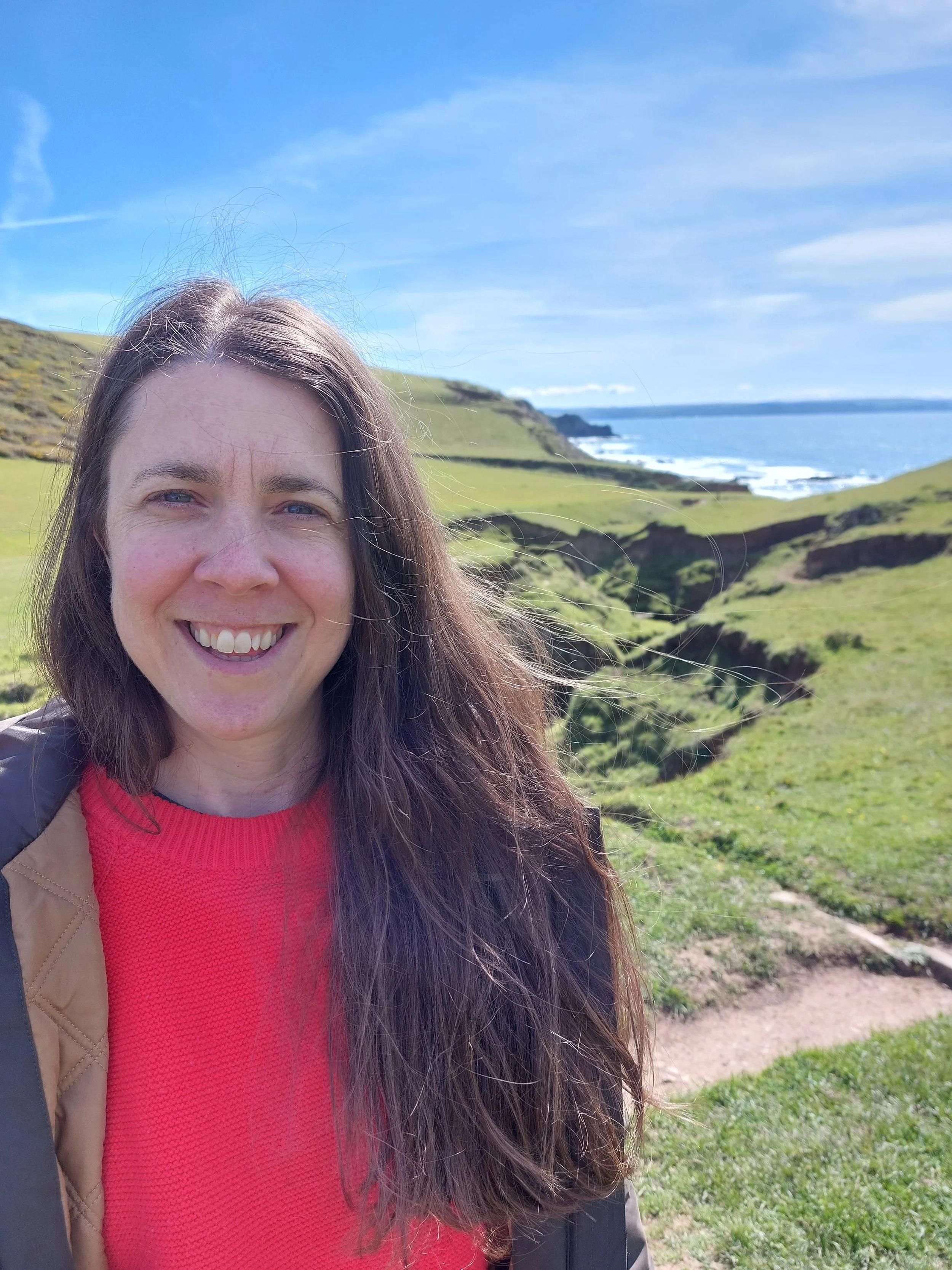 Lizzie Briggs smiling outdoors with green hills and ocean in the background under a partly cloudy sky.