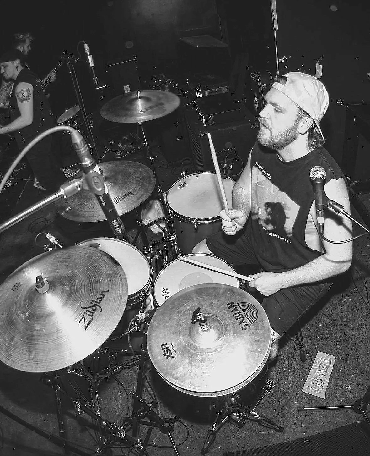 A man playing a drum set on stage, wearing a baseball cap backwards and a sleeveless shirt, with microphones and other band members in the background.
