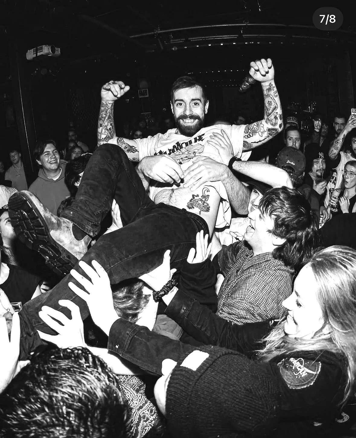 People at a concert crowd surfing with one person smiling and raising their arms while being held up by others.