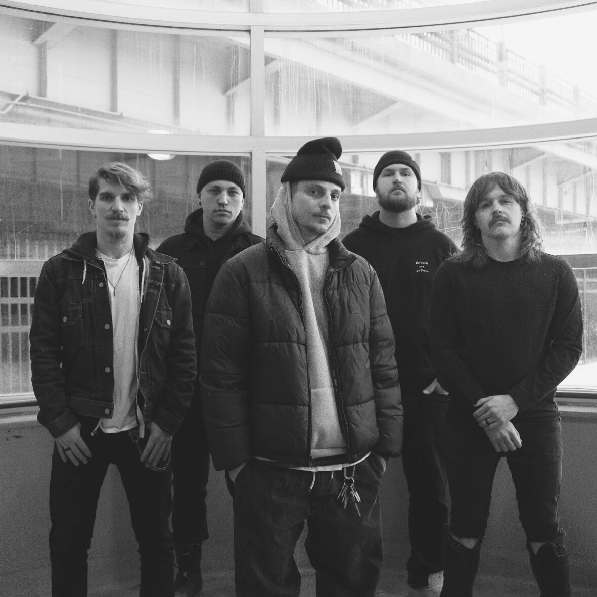 Black and white photo of five young men standing in front of a large glass window, dressed in casual, streetwear clothing including hoodies, jackets, and beanies.