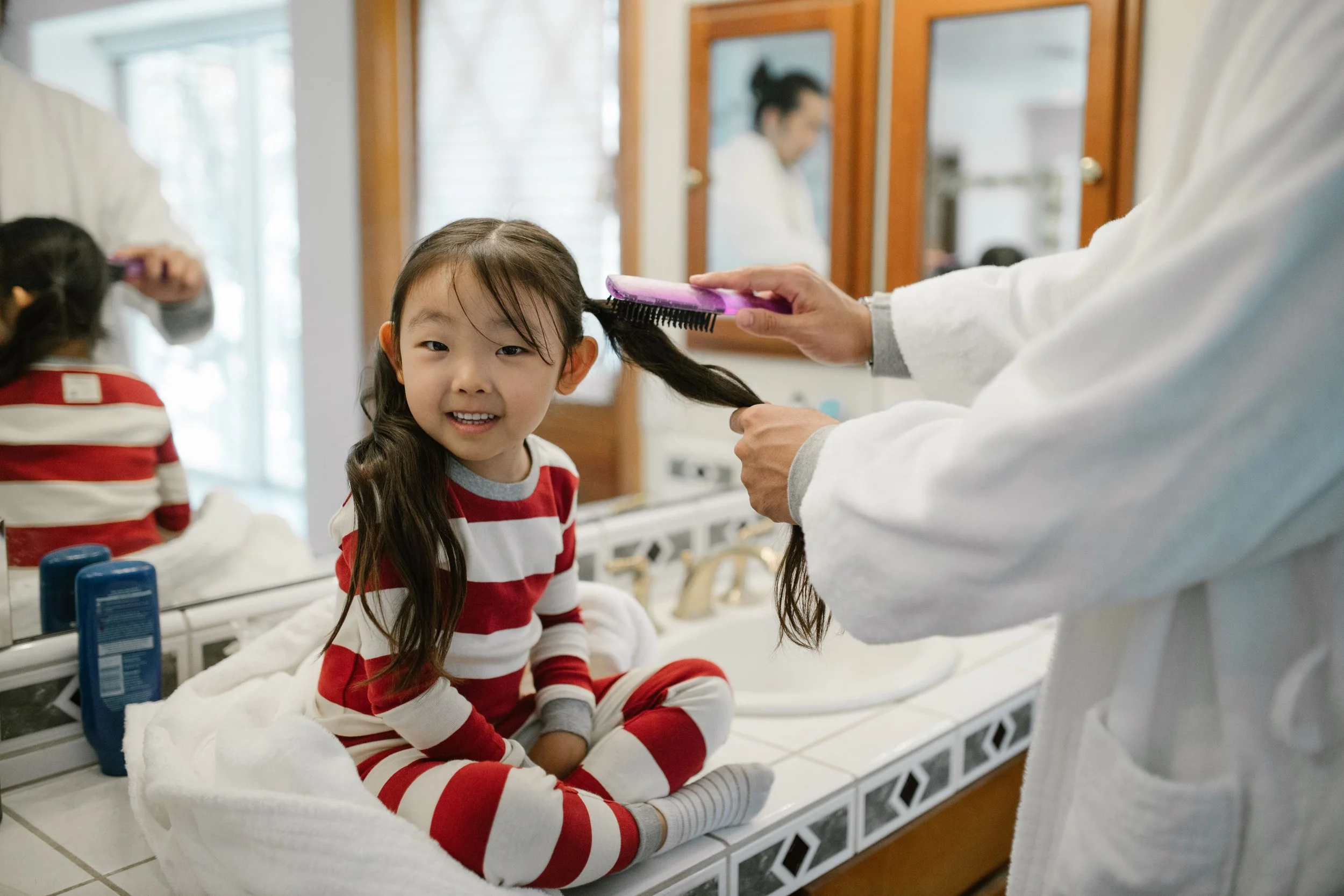 A young girl with long dark hair getting ready for school sitting on a white bathroom countertop in front of a mirror, getting her hair brushed by a person in a white coat. She is smiling and wearing a red and white striped shirt.