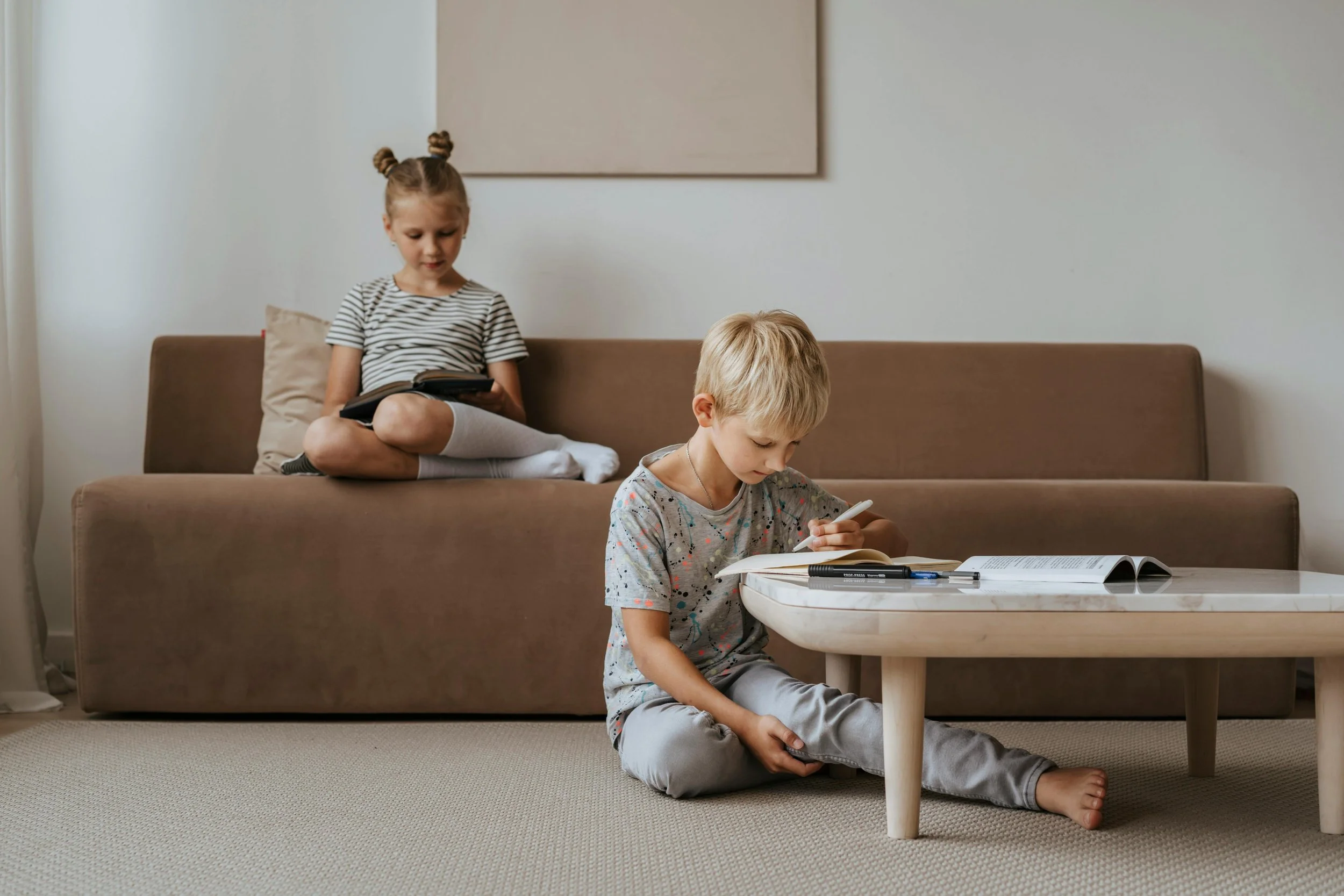 Two children reading at home; one girl sitting on a sofa and a boy sitting on the floor by a coffee table, both focused on their books.