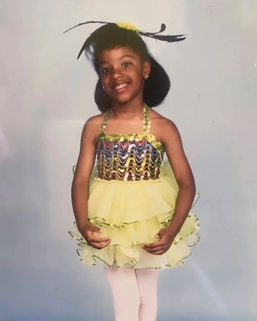 Young girl smiling, wearing a colorful dress with ruffled yellow skirt, white tights, and a large feathered hair accessory.