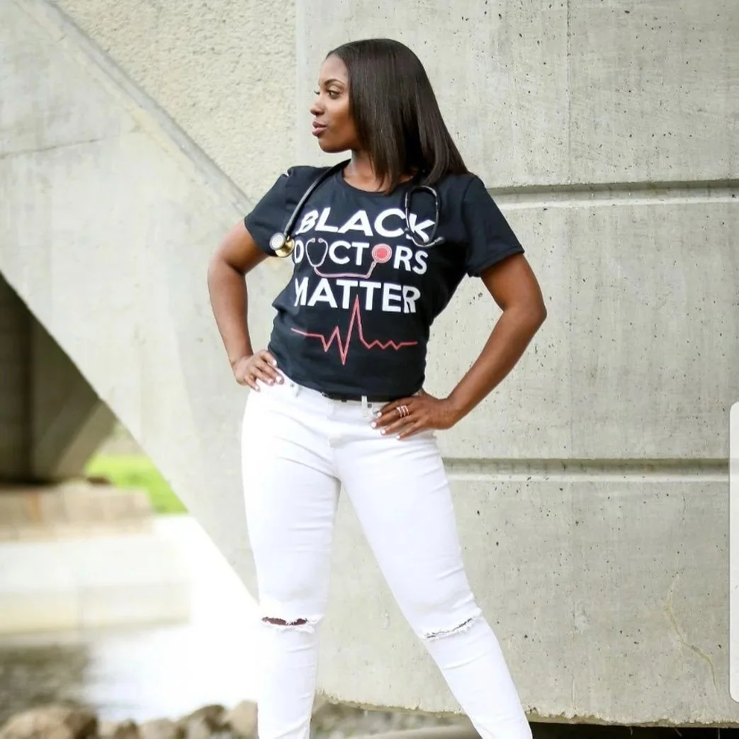 A woman standing outdoors, wearing a black T-shirt that says 'Black Doctors Matter' with a stethoscope graphic and a heartbeat line, and white ripped jeans. She has a stethoscope around her neck and her hands on her hips.