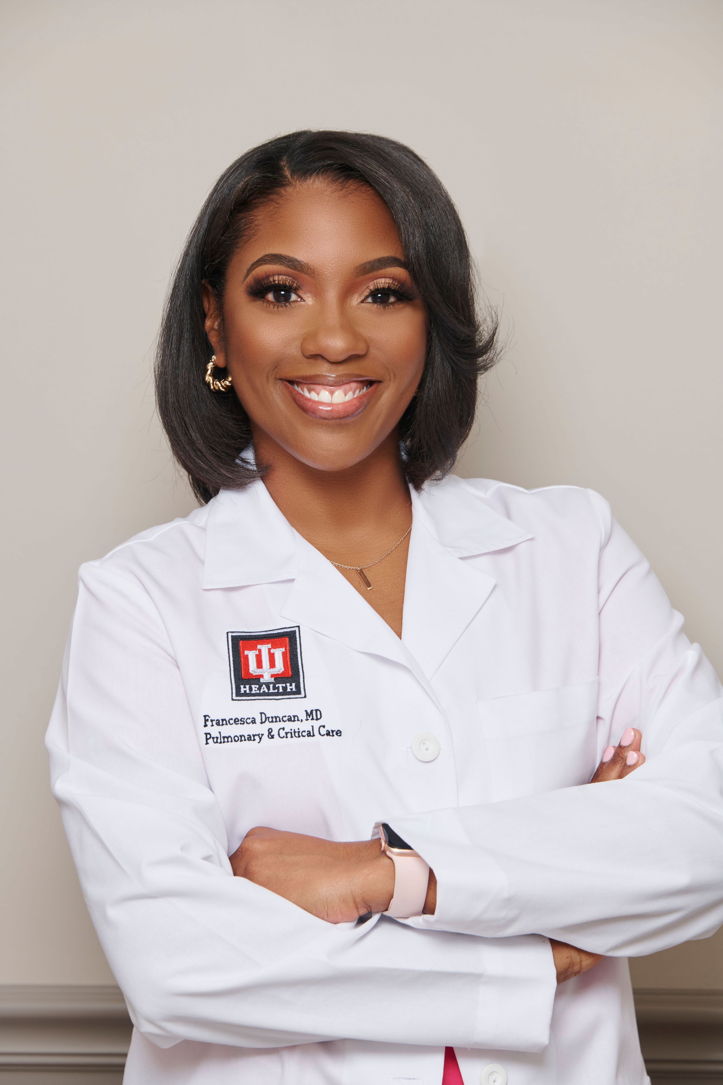 Portrait of a smiling African American woman with shoulder-length black hair, wearing a white medical coat with Indiana University Health logo, and crossed arms, standing against a plain light-colored wall.