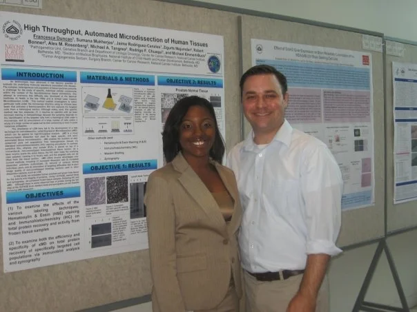 Two people, a woman and a man, standing in front of academic research posters at a conference or presentation event.