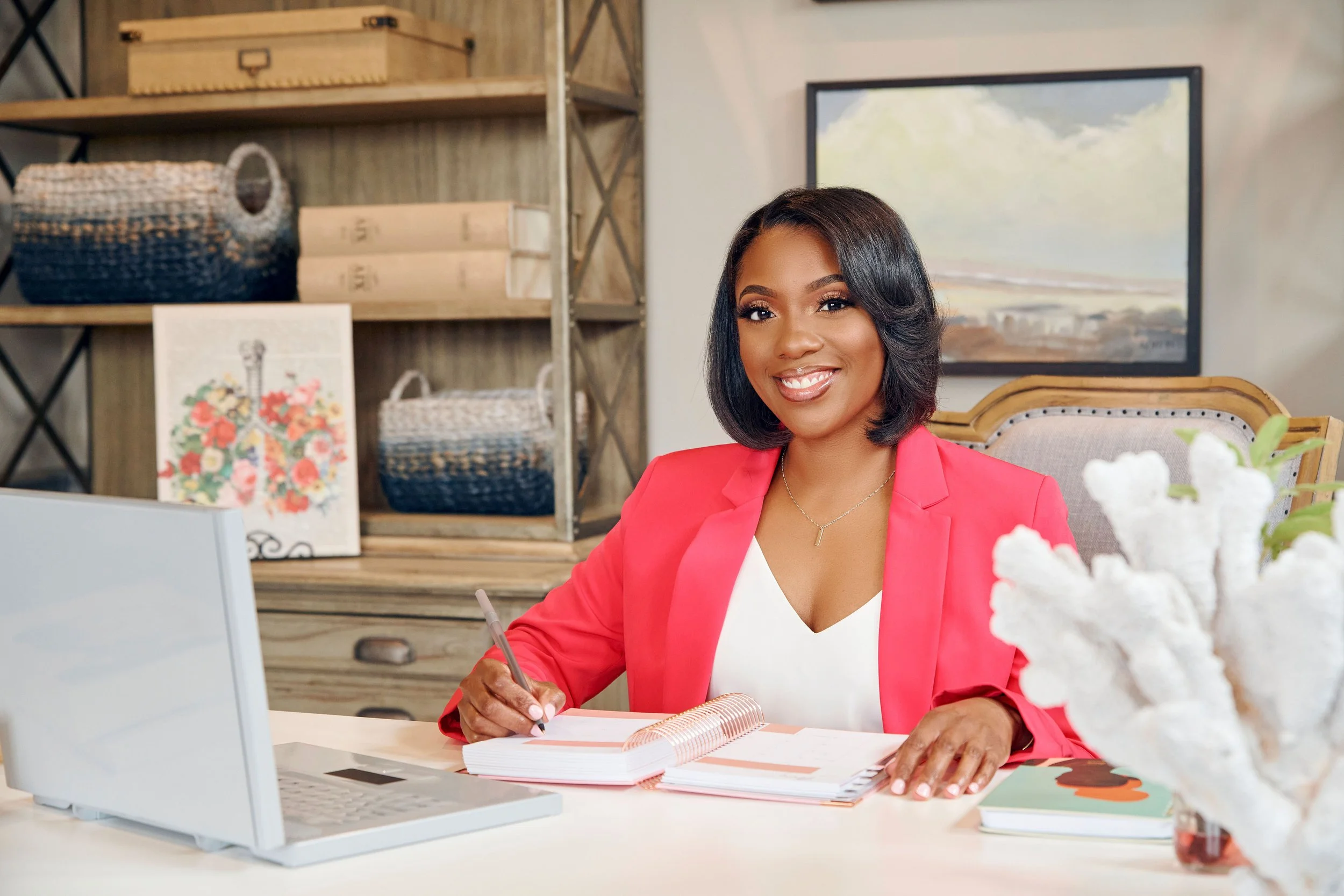 A smiling woman in a pink blazer sitting at a desk with a notebook and pen, in an office or studio setting with decorative baskets, artwork, and a landscape painting in the background.