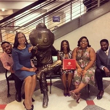 Five people sitting beside a large sculpture of a person's head and upper body, all smiling for the photo in a public indoor space.