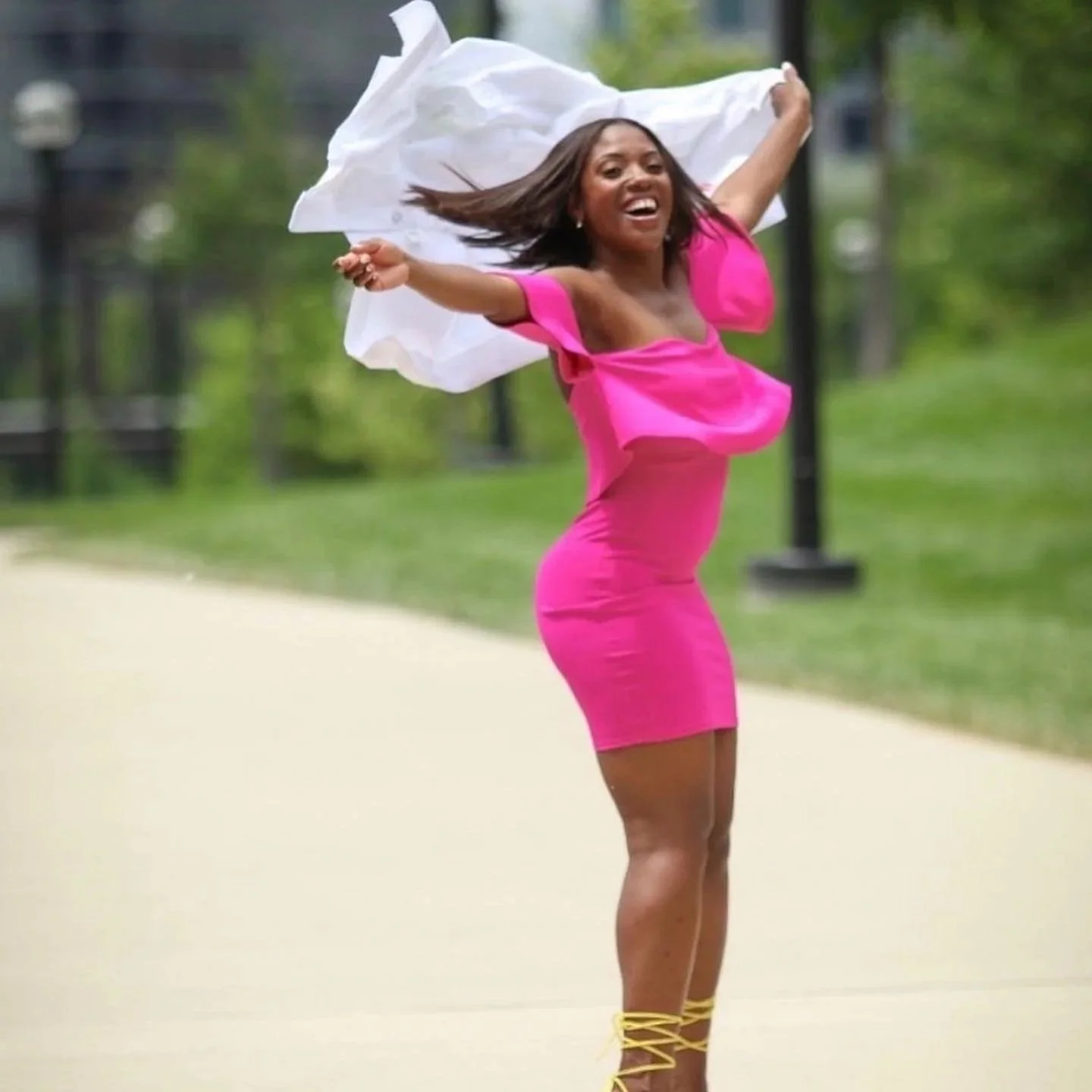 A woman in a bright pink dress joyfully spinning outdoors on a sunny day, holding a white cloth in her arms and smiling.