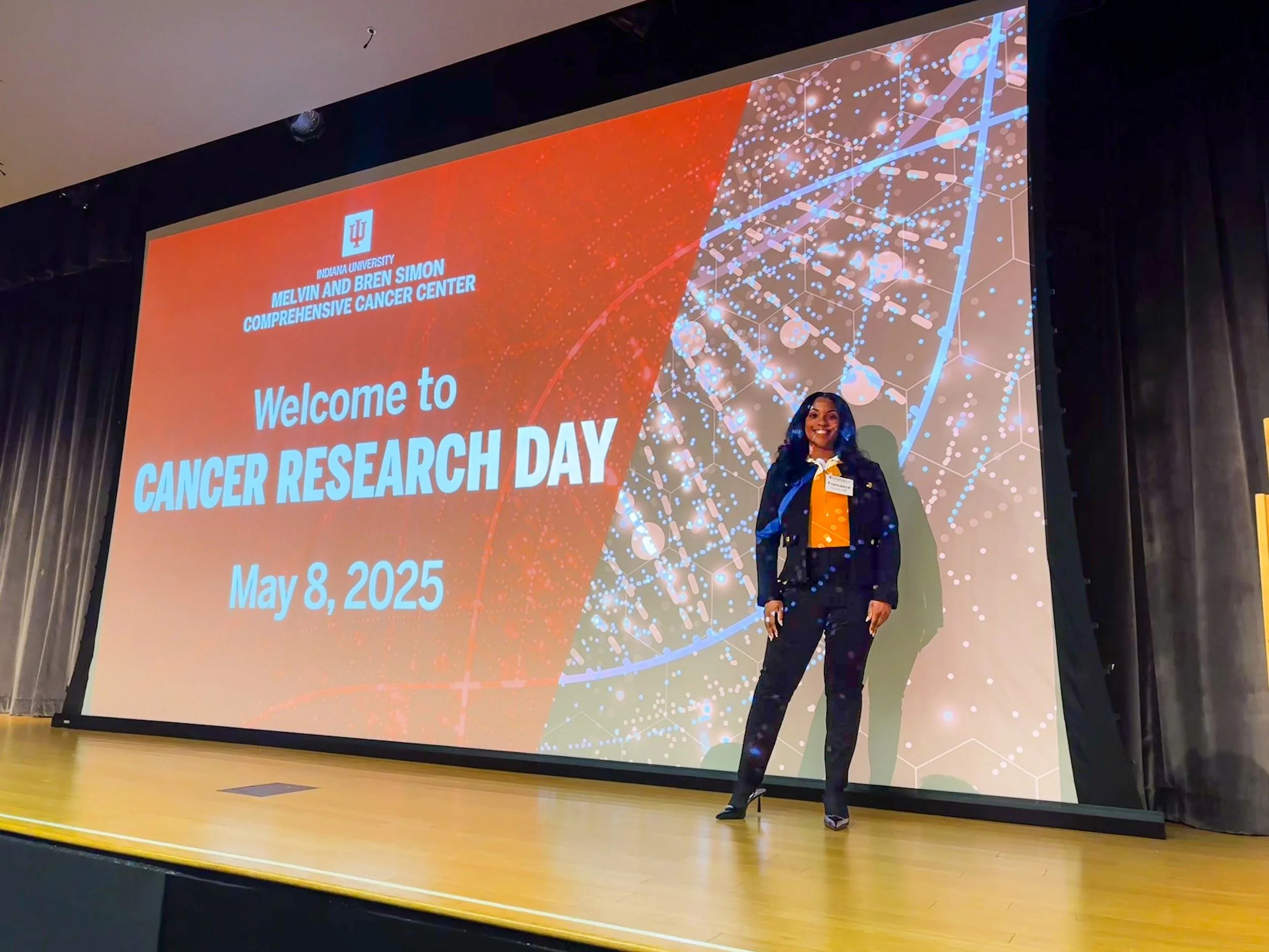 A woman standing on stage in front of a large presentation screen at an event called Cancer Research Day, dated May 8, 2025, hosted by Indiana University Melvin and Bren Simon Comprehensive Cancer Center. She is dressed in a dark blazer, white blouse, and high heels, smiling toward the camera.