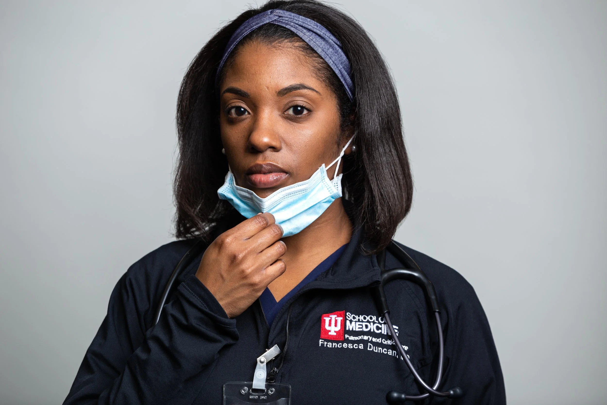 A woman wearing a dark medical uniform with a stethoscope around her neck adjusts a blue face mask. She has dark hair with a purple headband and is looking directly at the camera. The uniform has a logo that reads 'Indiana University School of Medicine' and includes her name, Francesca Duncan.