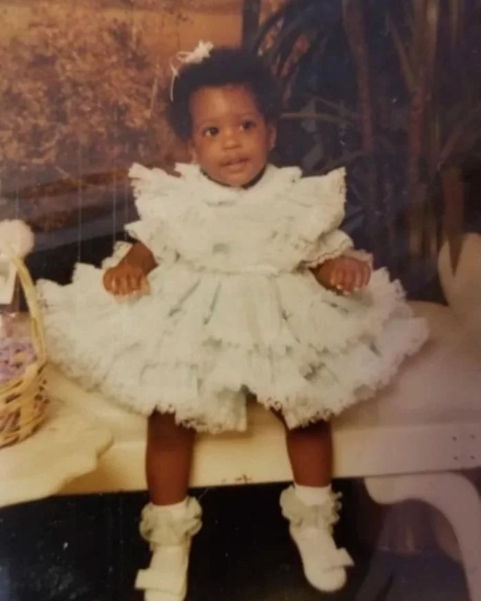 Young girl dressed in a white ruffled dress with puffed sleeves, wearing matching socks and shoes, sitting on a bench with a basket to her left and a plant behind her.