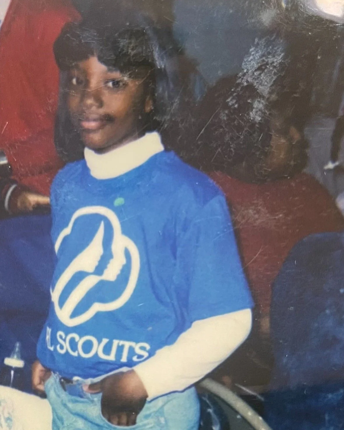 A young boy wearing a blue Scout uniform with a white collar and logo, standing indoors with other people around him.