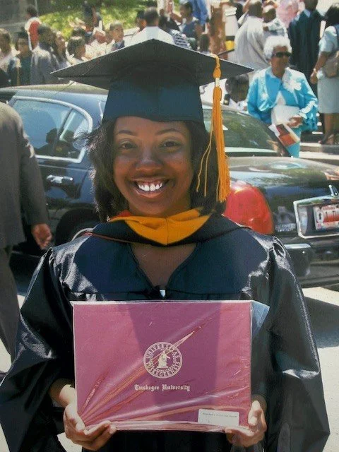 A young woman in a graduation cap and gown holding a diploma cover at a graduation ceremony outdoors.