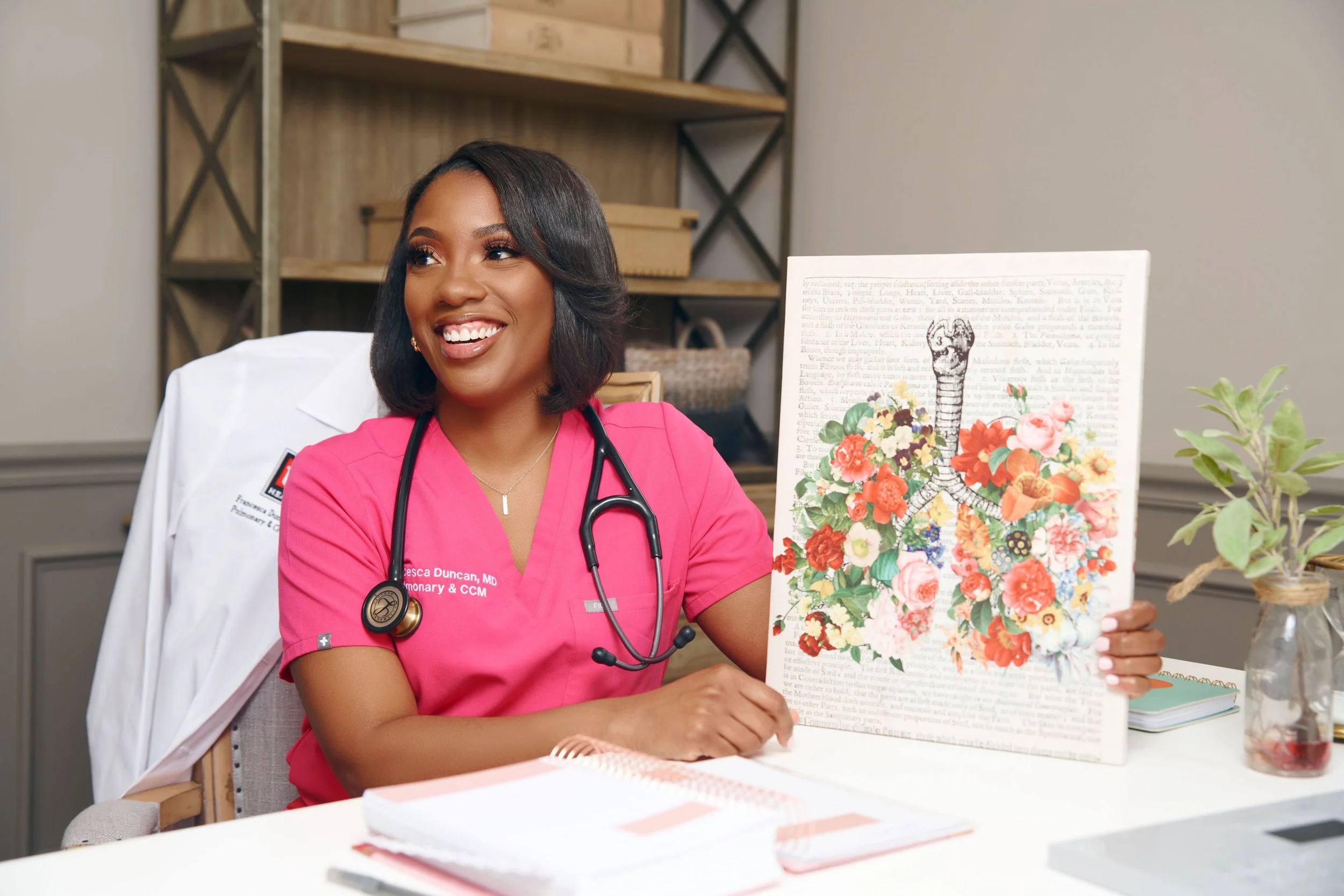 A smiling African American female doctor in pink scrubs with a stethoscope around her neck, sitting at a desk in a medical office, holding a colorful floral chart or artwork, with a plant on the desk and shelves in the background.