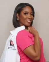 A smiling woman with dark hair, wearing a pink uniform and a white lab coat over her shoulder, standing against a plain background.