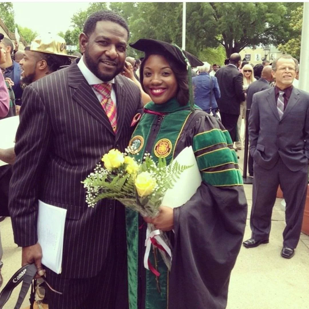 A man and woman smiling at a graduation ceremony, the woman in cap and gown holding a bouquet of yellow and white flowers.