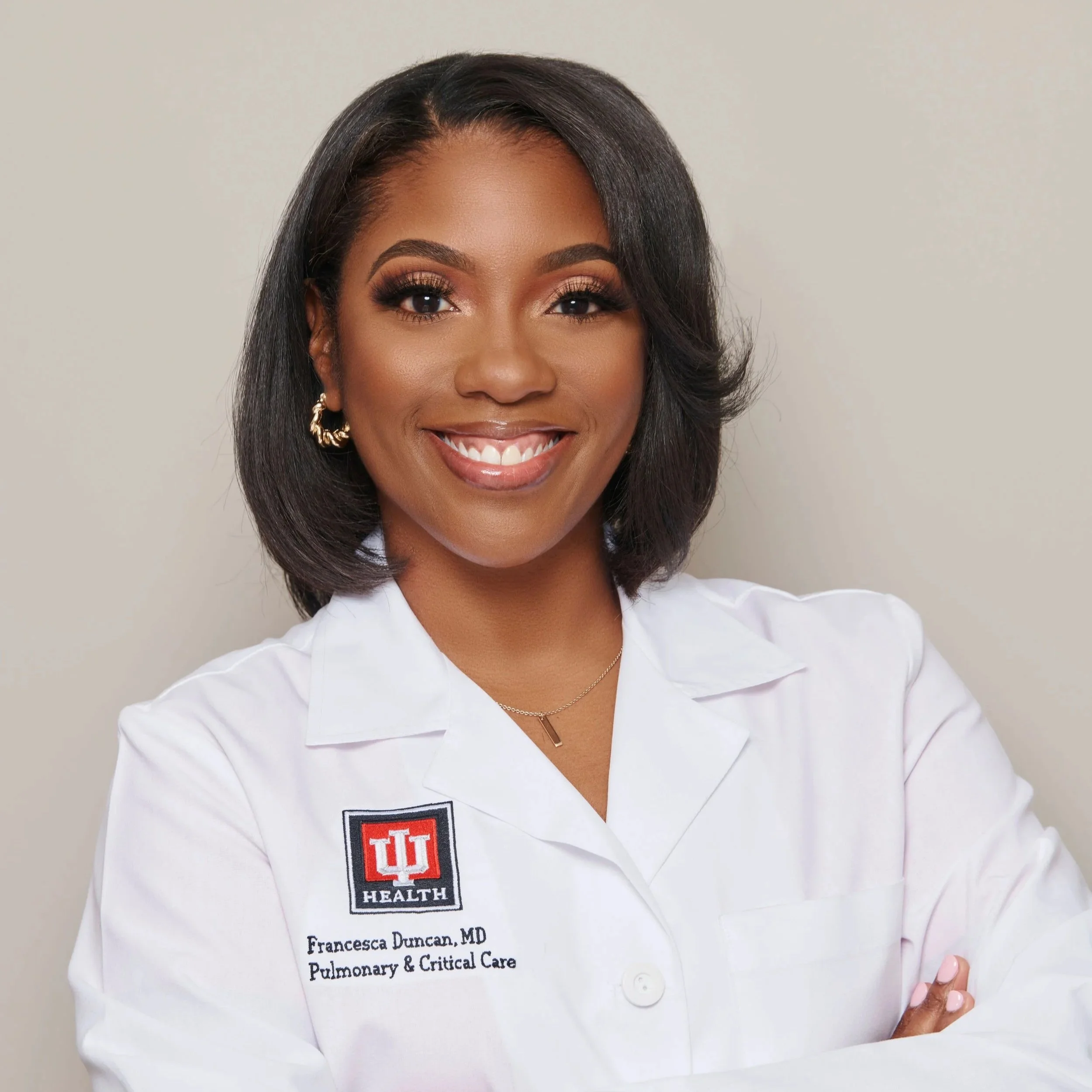Close-up of a woman wearing a white medical coat with an Indiana University Health badge, smiling, with shoulder-length black hair, light makeup, and gold hoop earrings.