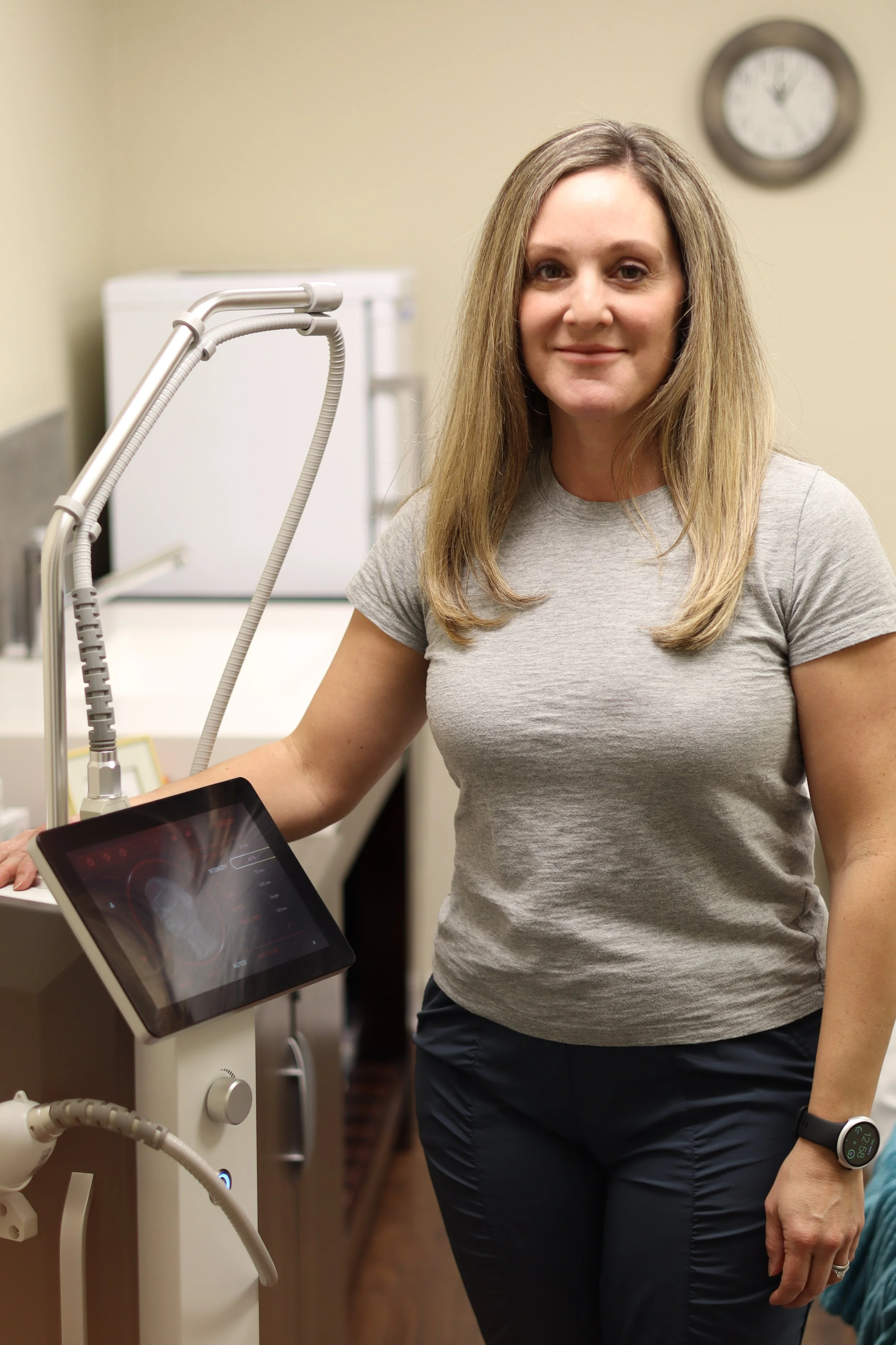 A woman in a gray t-shirt standing next to medical equipment with a tablet screen, in a clinical setting.
