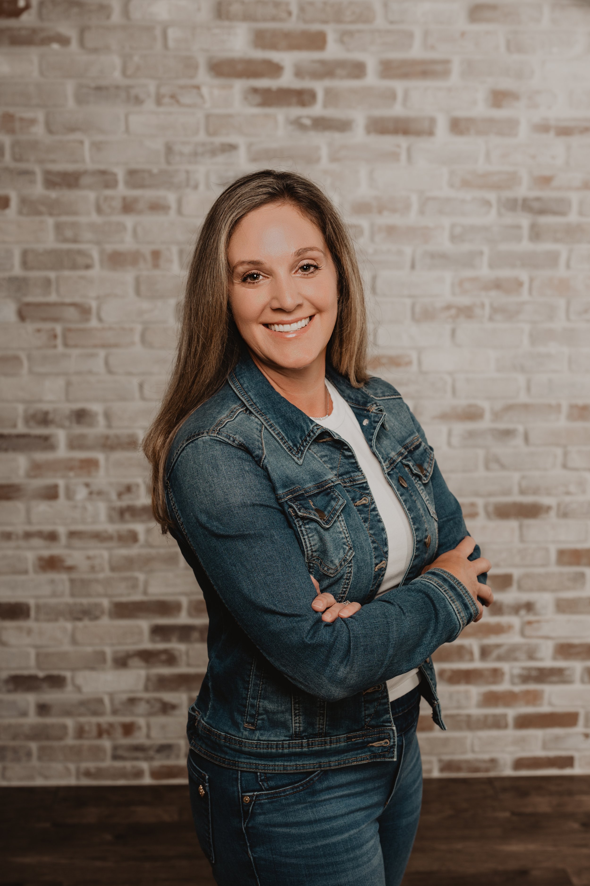 A woman standing with arms crossed, smiling, in front of a light-colored brick wall, wearing a denim jacket and jeans.
