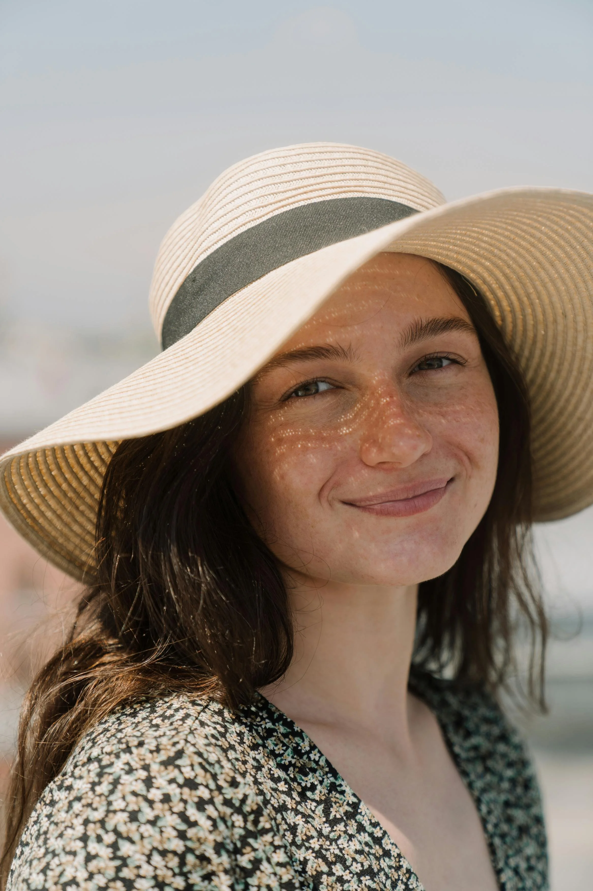 A woman with long dark hair, freckles, and a light smile wearing a wide-brimmed straw hat with a dark band and a black dress with white floral patterns, standing outdoors on a sunny day.