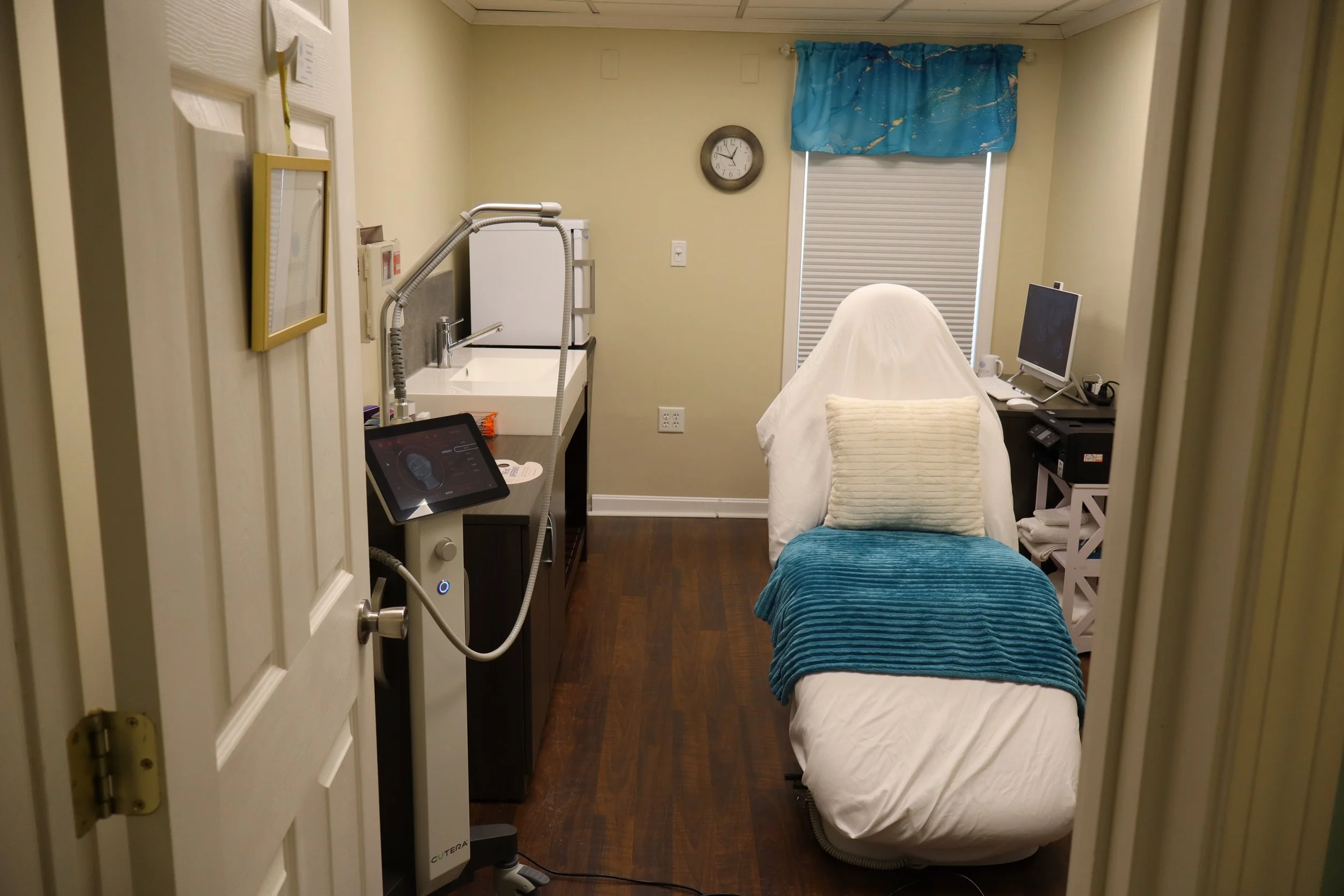 A medical examination room with a treatment bed covered in a white sheet and a teal blanket, a computer monitor on a side table, a blue window curtain, and medical equipment including a sink, tablet, and other devices.