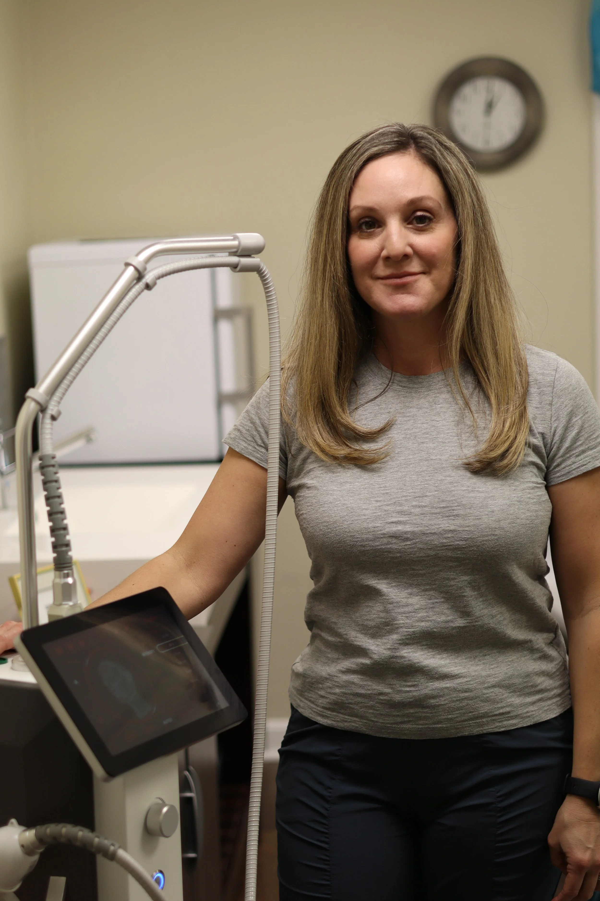 A woman with shoulder-length brown hair wearing a gray t-shirt standing in a medical or clinic setting next to a medical device with a display screen and a flexible arm.