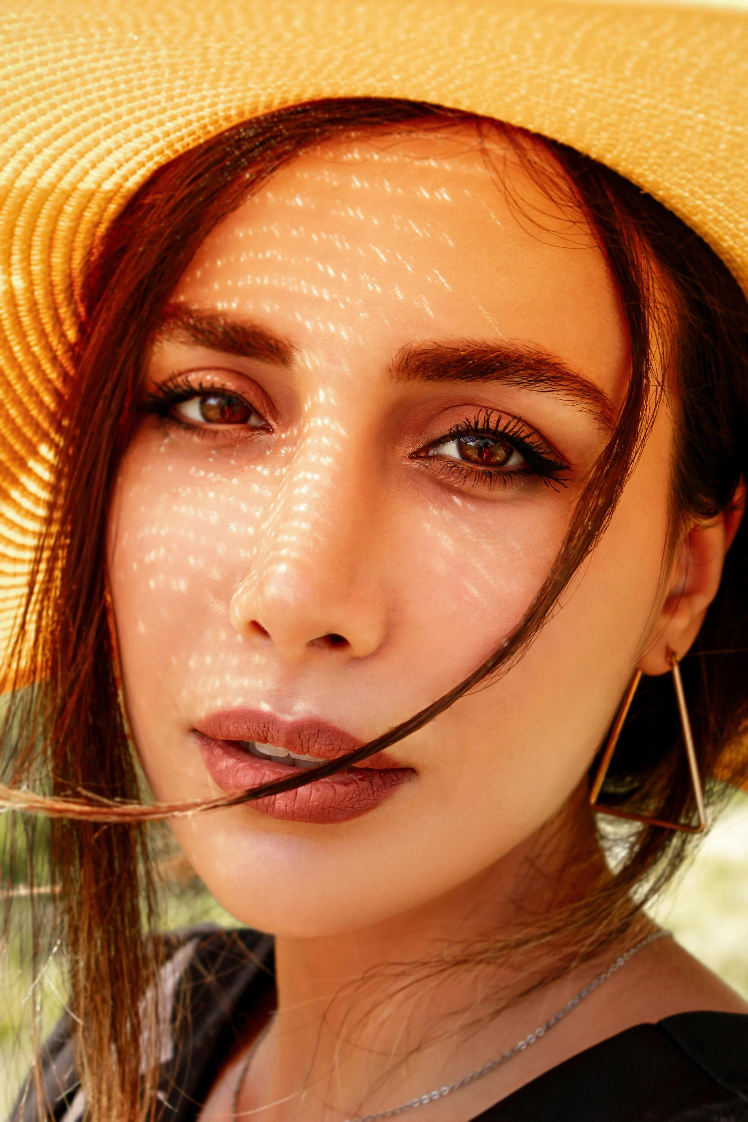 Close-up portrait of a woman wearing a yellow sunhat, with sunlight creating a dappled pattern on her face, and wearing a black top and geometric earrings.