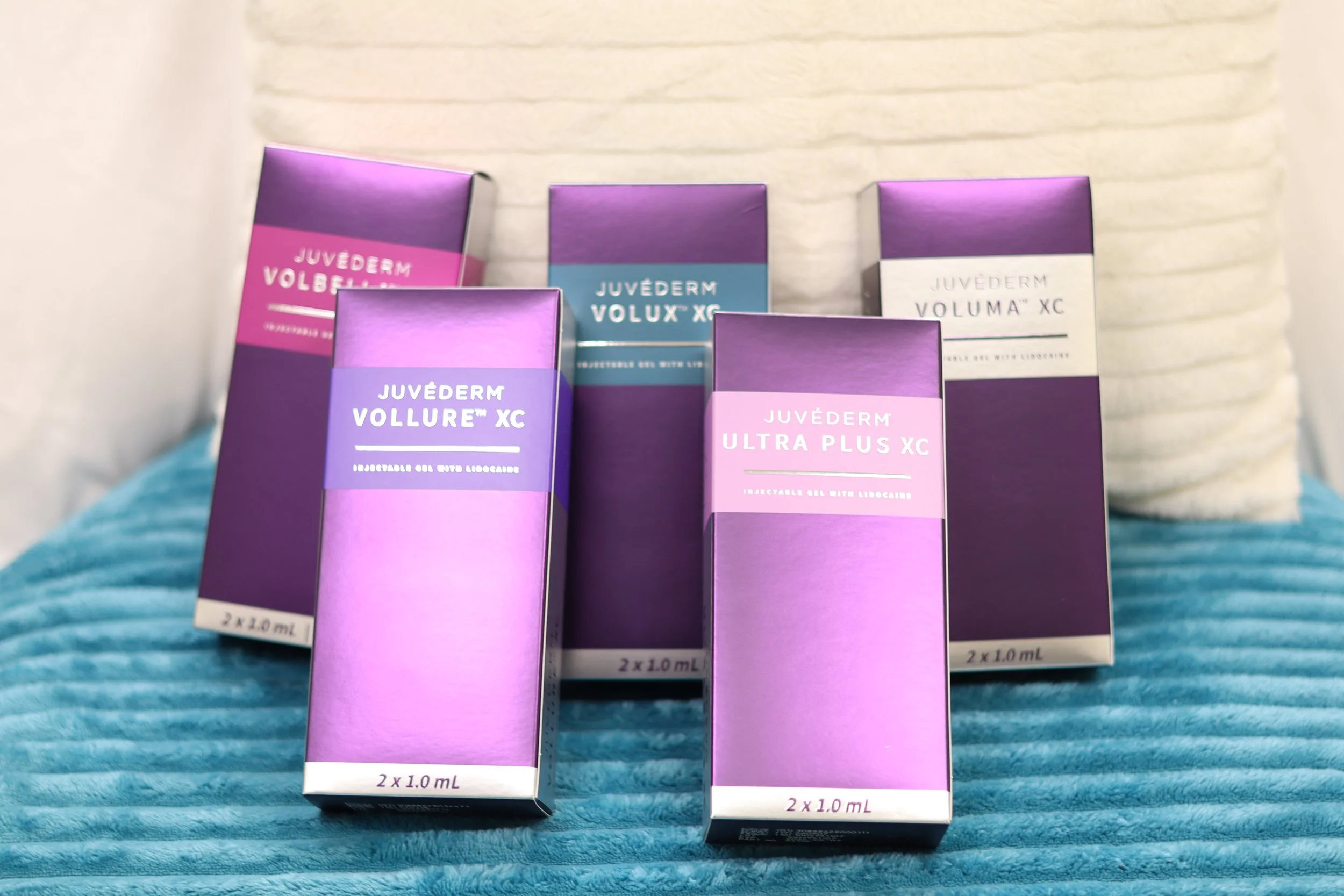 Five boxes of Juvederm dermal fillers are displayed on a blue textured surface, with a lightly textured white background. The boxes are purple with varying shades and white text, indicating different Juvederm products.