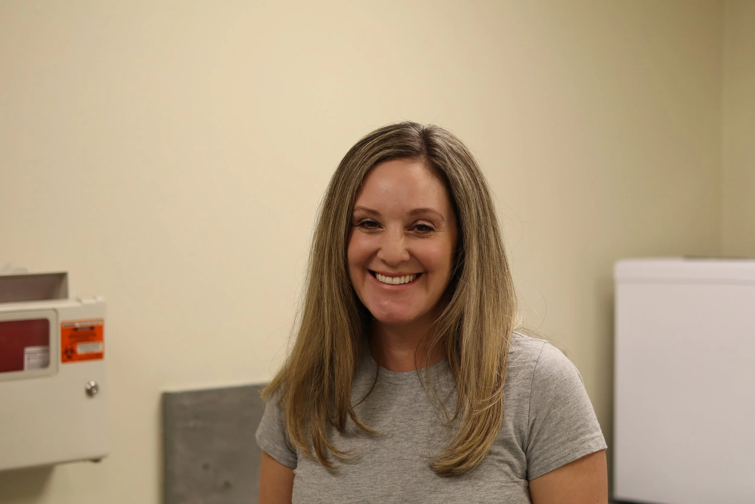 A woman with long blonde hair smiling in a medical or clinical setting, wearing a gray t-shirt, with a faint background of beige walls and medical equipment.