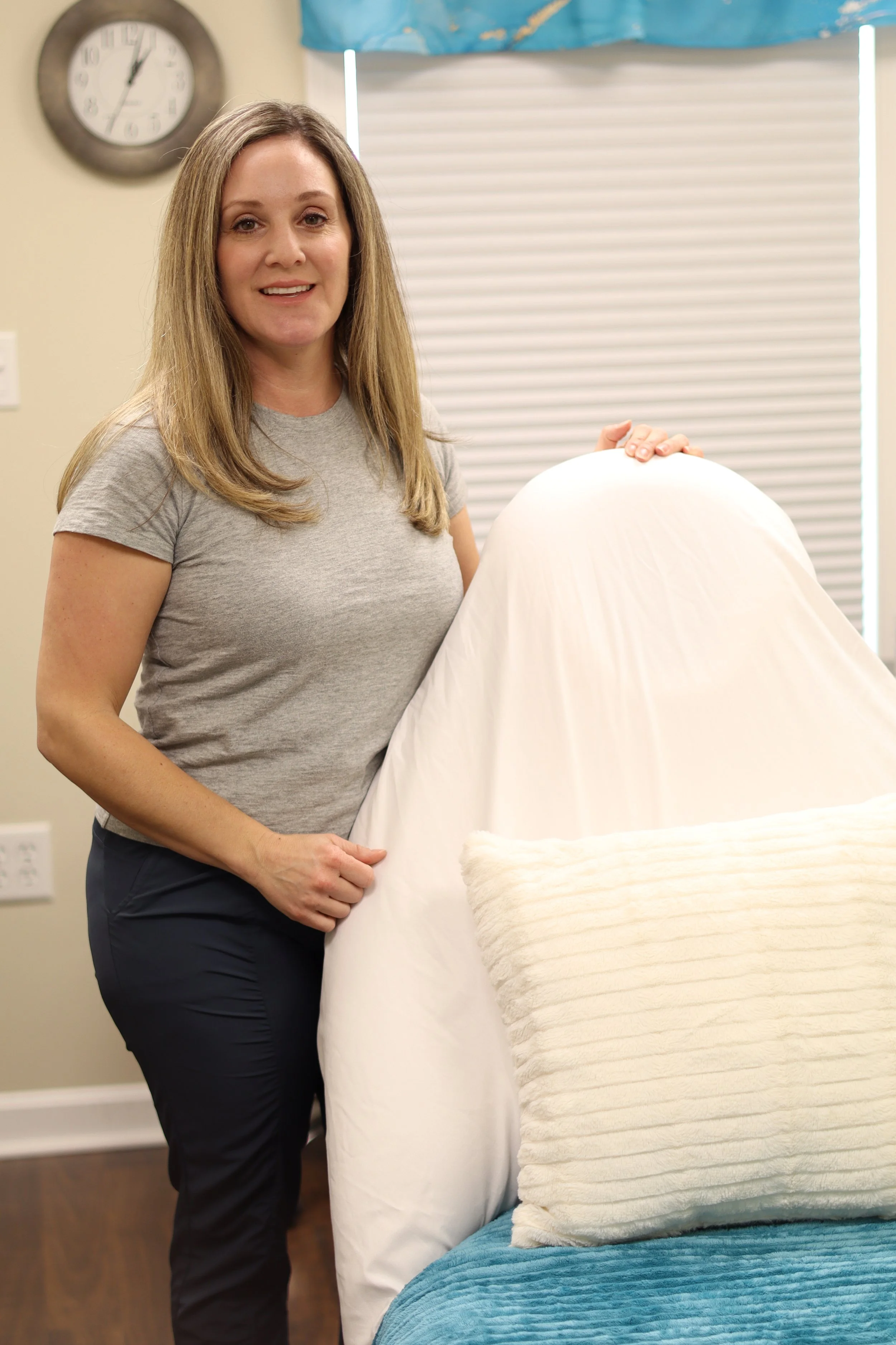 A woman with long blonde hair wearing a gray t-shirt and dark pants standing beside a bed with white sheets and a beige pillow, in a room with a clock on the wall and closed blinds.