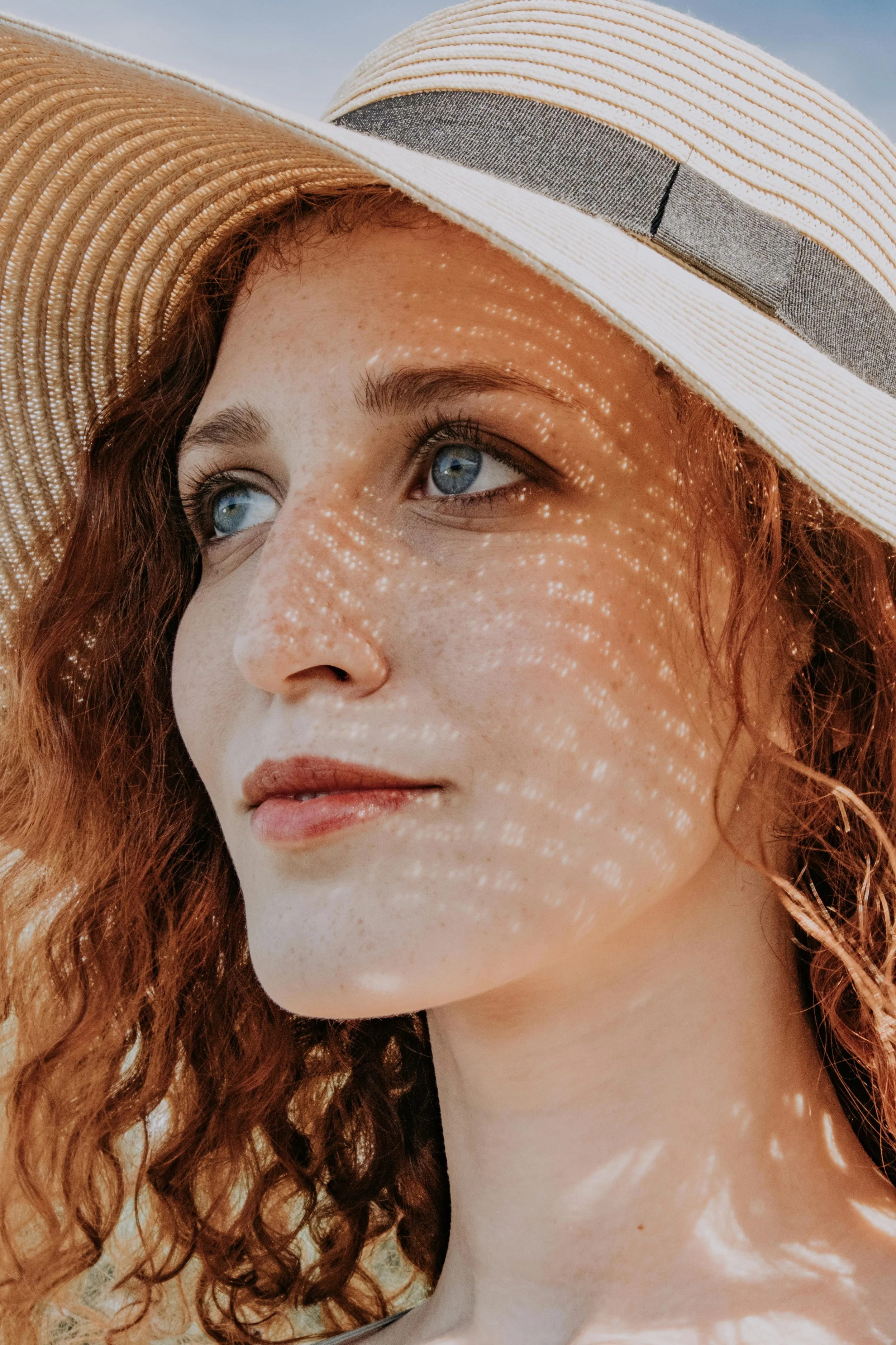 Close-up of a woman with curly red hair wearing a wide-brimmed straw hat, with sunlight creating patterned shadows on her face.