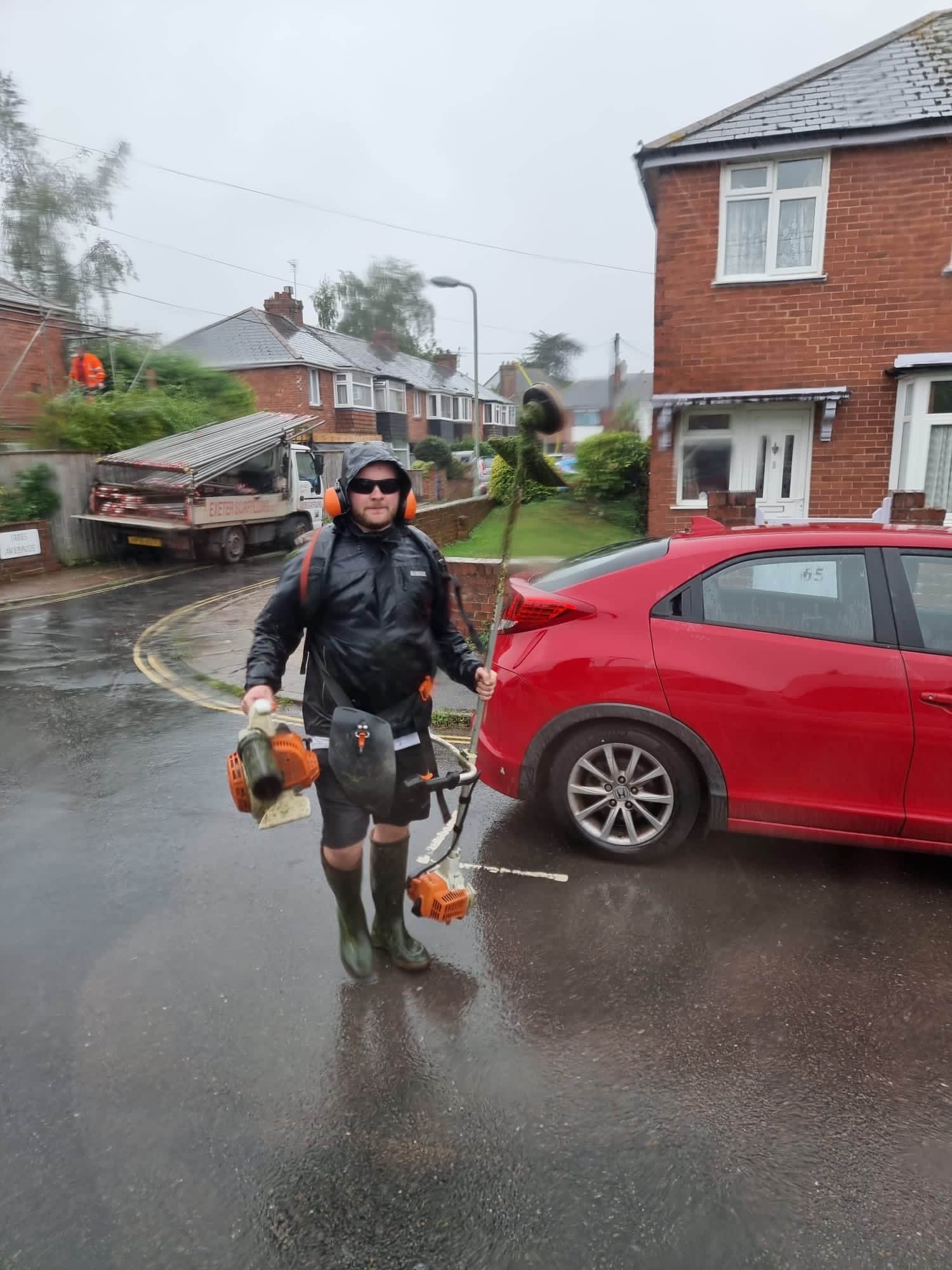 Man wearing rain gear and carrying outdoor equipment standing on a rainy residential street.