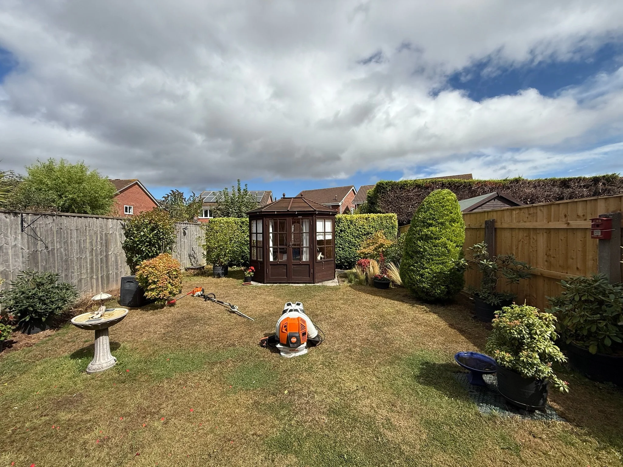A backyard garden with a grassy lawn, potted plants, a wooden fence, and a small glass gazebo against a partly cloudy sky.