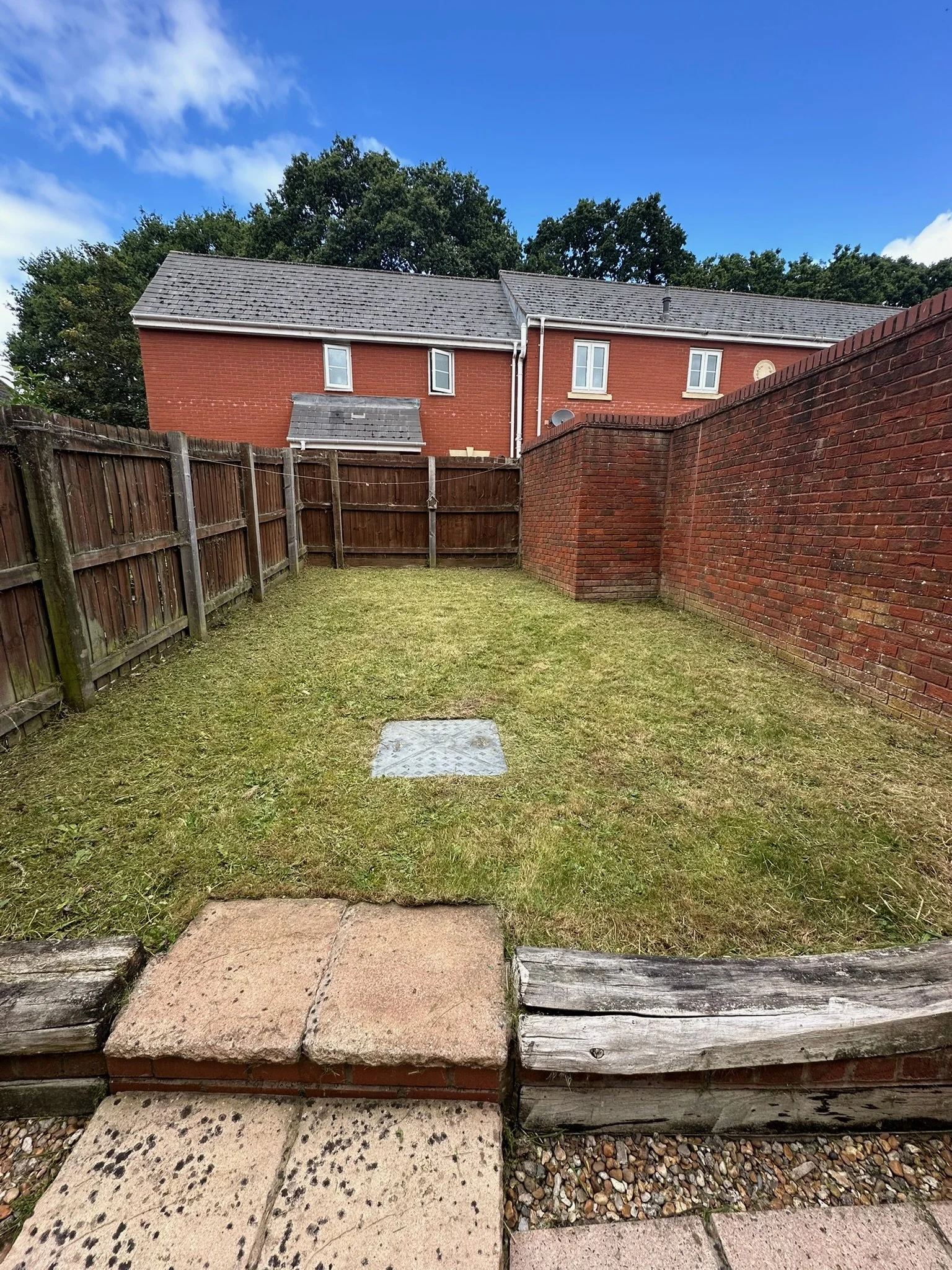 A small backyard with a grassy area, surrounded by wooden and brick fences, with a house with red brick walls and a gray tiled roof in the background.