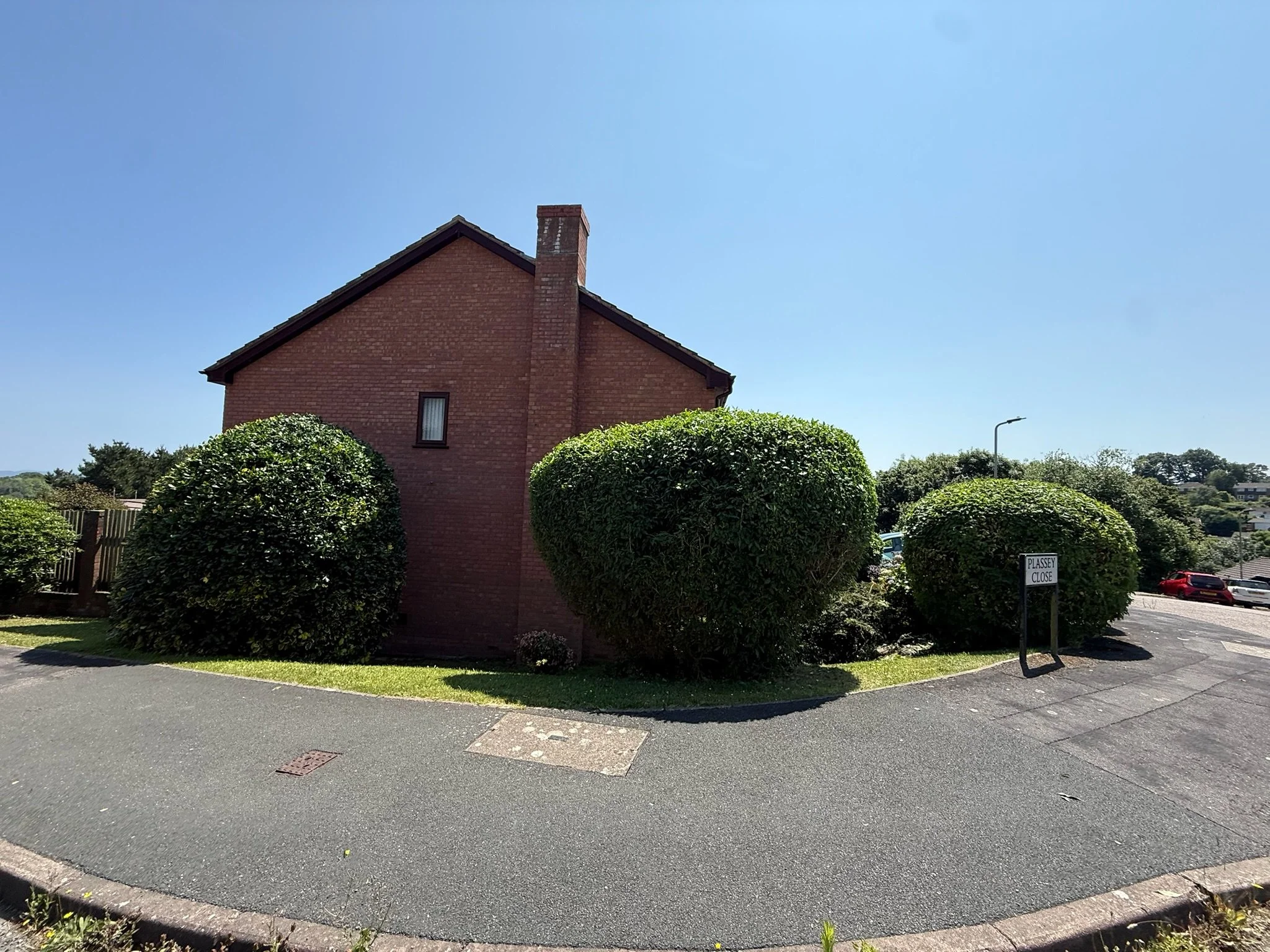 A red brick house with a sloped roof and a chimney, surrounded by large, neatly trimmed bushes, on a sunny day with a clear blue sky.