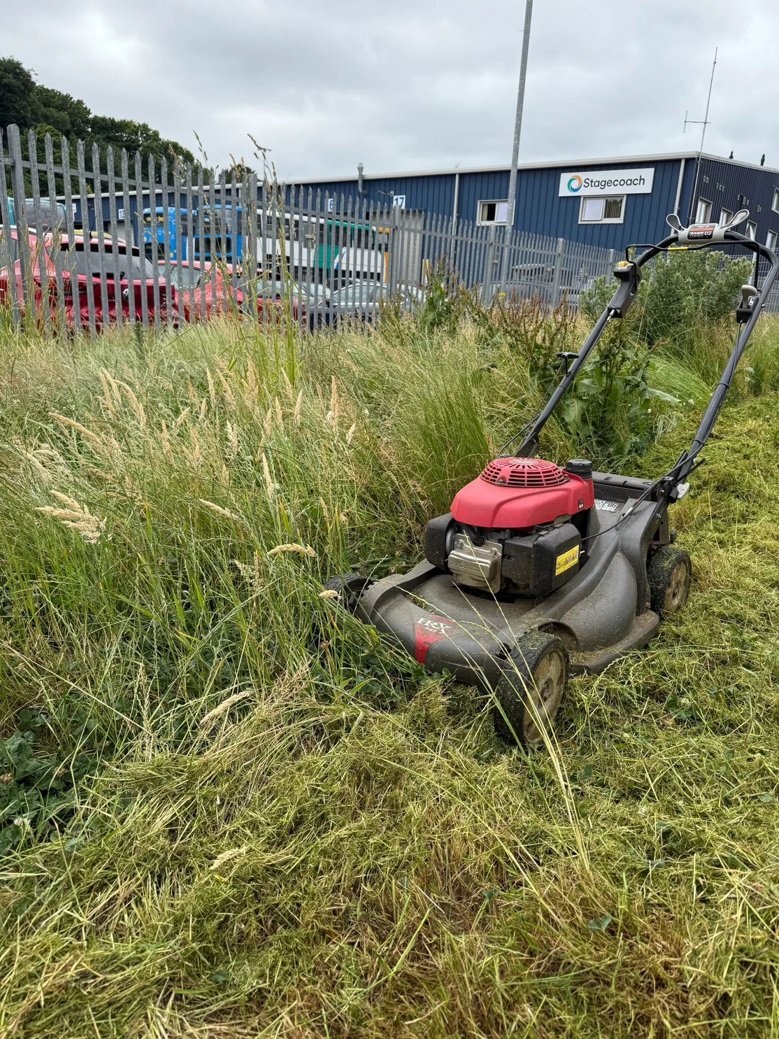 A lawn mower on overgrown grass in a grassy area outside a parking lot with a blue fence and a building labeled 'Stagecoach' in the background.