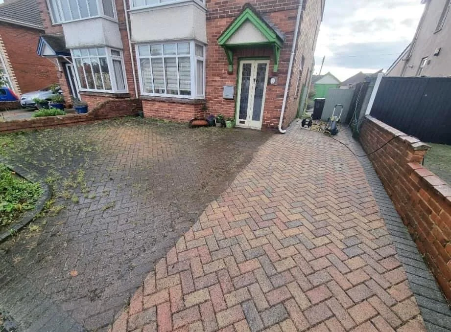 Driveway with half water and half dry brick pavement leading to house entrance, with garden beds, potted plants, a black fence, and a cloudy sky.