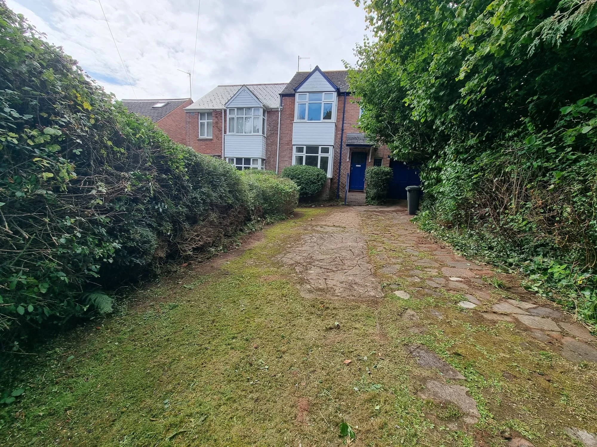 View of a residential driveway leading to a brick house with white bay windows, surrounded by bushes and trees, with a grassy and cobblestone surface.
