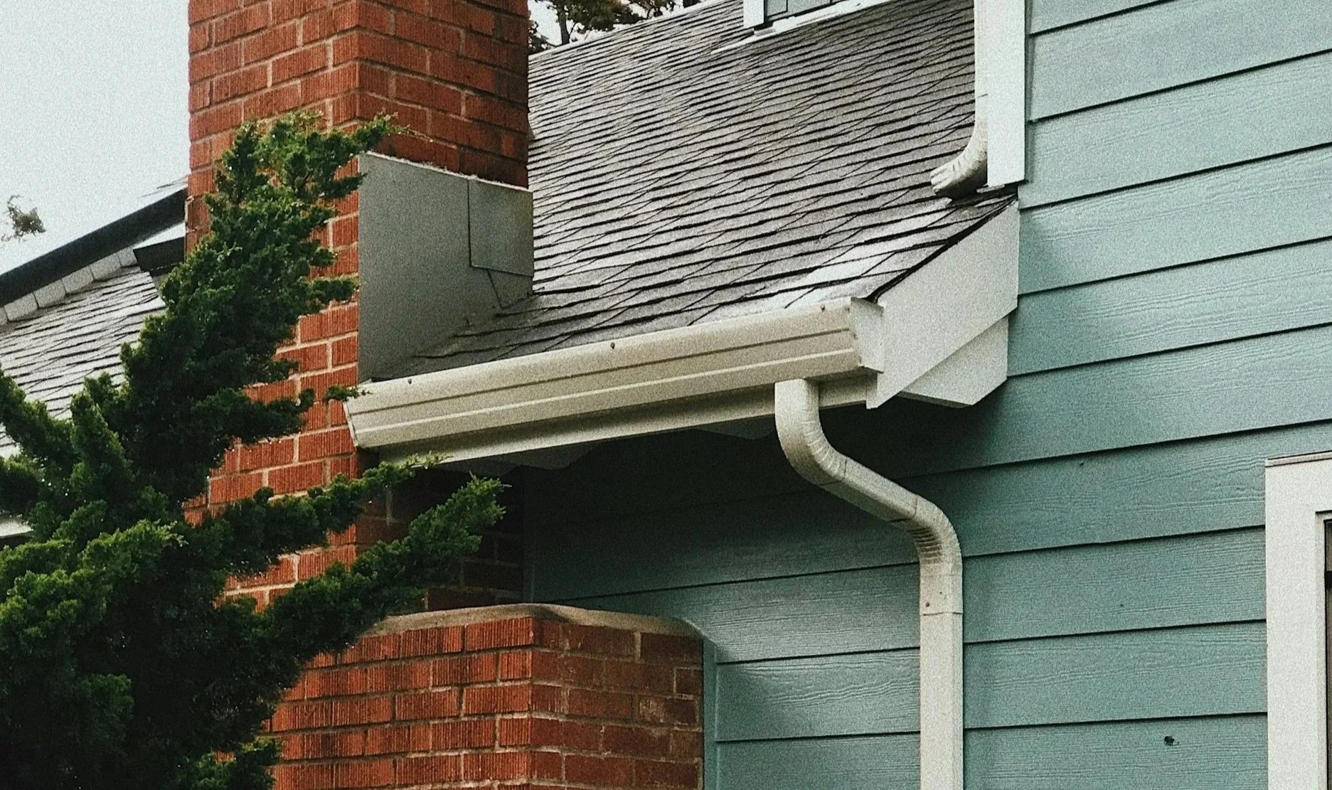 Close-up of a house corner showing a brick chimney, blue wooden siding, a gray shingled roof, and a white gutter system with a downspout, partially obscured by a green bush.