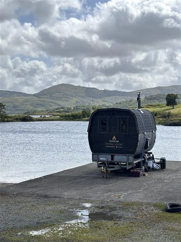 Small black sauna located near a lake with green hills and cloudy sky in the background.