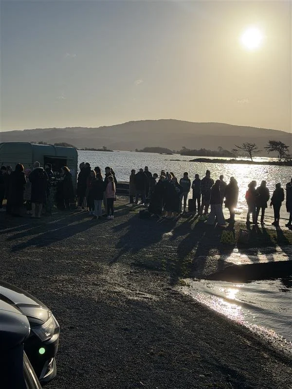 Group of people gathered by a river at sunset, with hills in the background and cars parked nearby.
