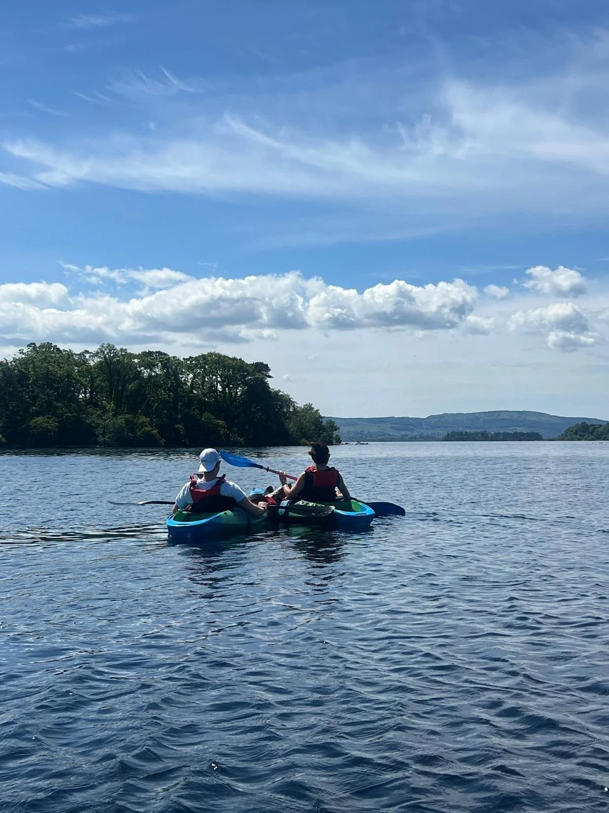 Two people kayaking on a lake with clear blue sky and some clouds, surrounded by trees and distant hills.