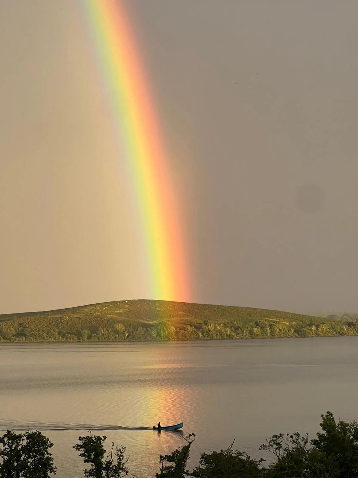 A rainbow over a lake with a boat and a person inside, a island or hill in the background, and trees in the foreground.